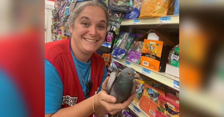 a woman smiling as she holds bob the pigeon in her hands. she's standing next to shelves of bird food in a store.