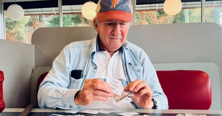 man sitting in a waffle house booth. he's wearing a hat and is holding onto a five dollar bill and a note that says "love every body." cash of various amounts is spread out on the table along with notes with that same message.