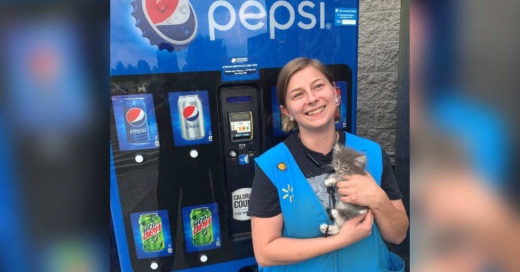 lindsey russell wearing her walmart uniform as she stands in front of a pepsi vending machine. she's smiling as she holds the small, grey kitten that was rescued from inside the vending machine.