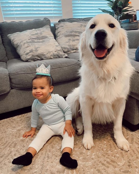 vanora sitting on the floor and smiling as she wears a birthday crown that says "one." she is sitting next to taco the golden retriever who is smiling as well. 
