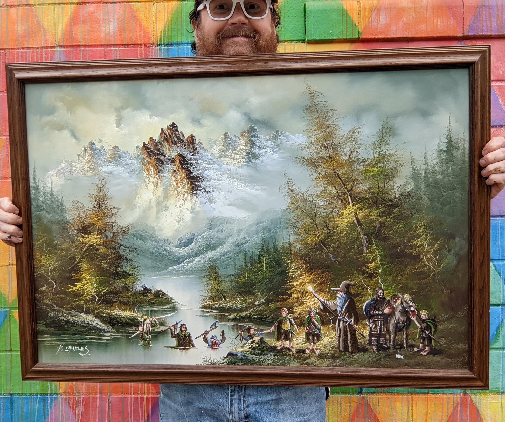 a man with a beard and glasses smiling as he holds a large painting in front of him that covers up most of him. the painting has lots of trees, a river, and a large, snowy mountain in the distance. "lord of the ring" characters have been added and are making their way across the river in a   single file line.