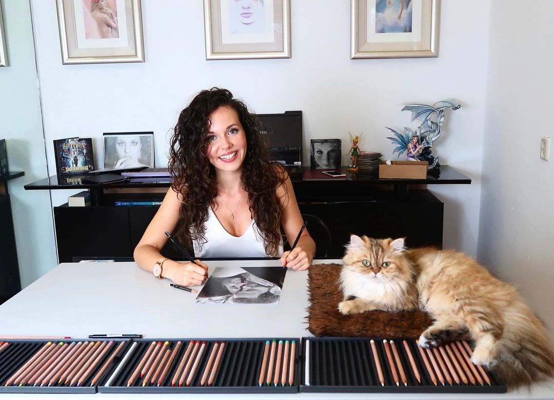 Dutch artist Rajacenna van Dam smiles while sitting at her desk drawing with her cat.
