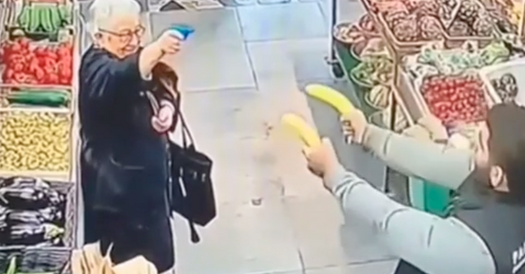elderly woman pointing at a grocery store employee with a small toy gun and the grocery store employee is pointing two bananas at the elderly woman in the middle of the produce section.