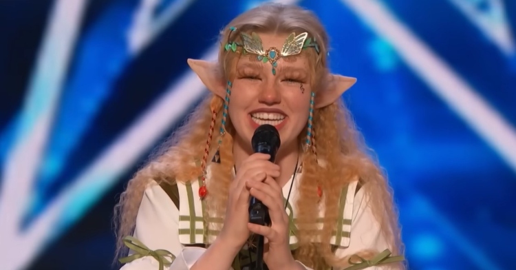 freckled zelda smiling as she holds a microphone and sings on “america’s got talent.” she’s wearing a fairy outfit that includes a headpiece and elf ears.