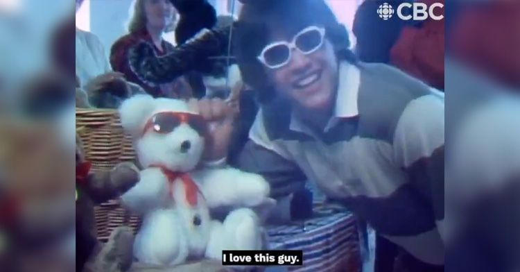 keanu reeves giving a news report in 1984. he’s wearing white sunglasses and smiling as he looks at the camera. he’s leaning down toward a white teddy bear in red sunglasses and is holding onto one of its ears. keanu reeves is captioned saying about the bear, “I love this guy.”