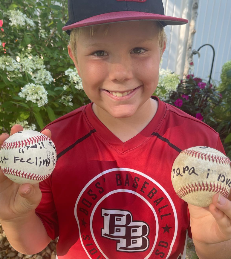 felix wearing his baseball uniform and holding his two home run balls that he signed. one of them has his name and says "best feeling ever" and another one says "papa i love you."
