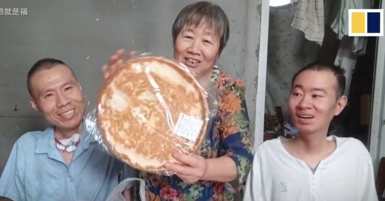 mama lu smiling as she stands between her two songs. she’s holding up a round, flat bread. xiaoqiang and xiaomeng are on either side of her, smiling as well.