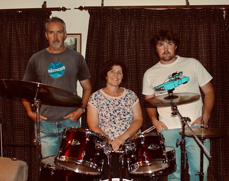 ryan stokes, his dad, and his mom, andrea, posing in their living room with curtains as the background. ryan and dad are standing and have serious looks on their faces as andrea sits at her drum set, smile on her face.
