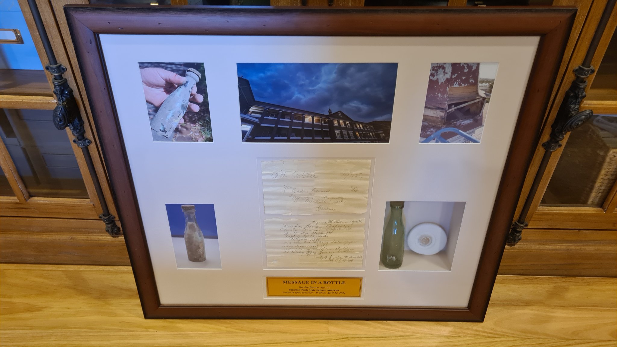 a display case featuring gordon's note from 1935 in the middle. above it is a photo of the school. to the right is the bottle and a photo of the spire where the note came from. to the left of the letter is a closeup photo of the bottle and a photo of the bottle sat on a table.