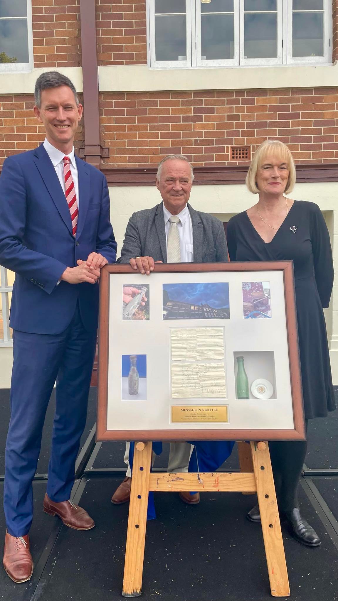 mark bailey, transport minister of australia, smiling as he poses with geoffrey benson and marilyn blundell. they're all standing behind the display cases featuring their dad's letter and bottle.
