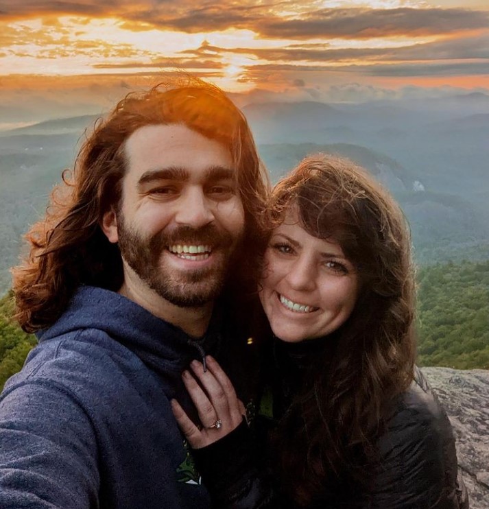 andrew renfro and abby powell smiling for a selfie together on top of a mountain with the sunset in the background. abby is wearing the engagement ring she just got after andrew proposed.