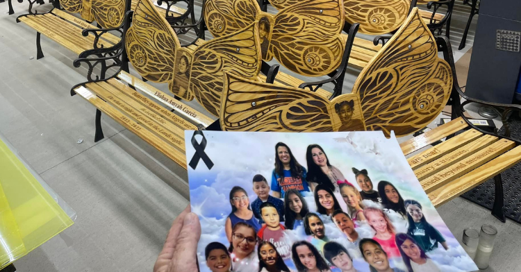 a hand holding a picture of uvalde shooting victims and the memorial handmade benches in the background