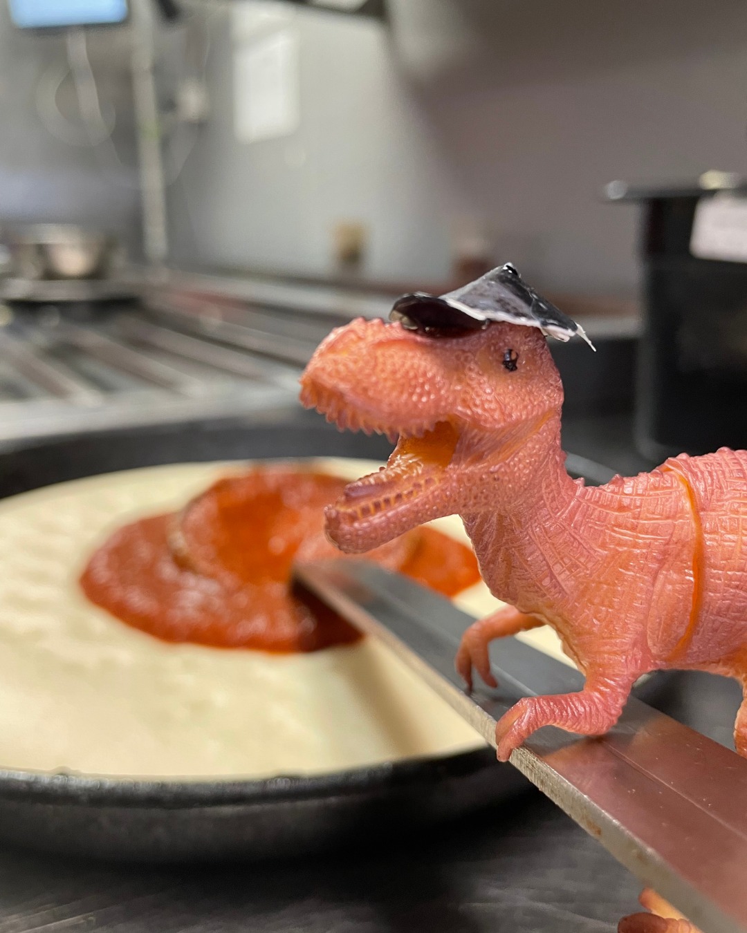 Bitey the dinosaur toy making pizza at Domino's. He's "holding" a utensil to spread sauce on a pizza.