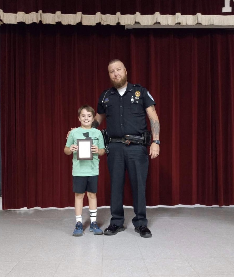 Kingston Police Department awards Gavin Keeney with hero award. Gavin smiles as he holds his award and an officer poses with him.