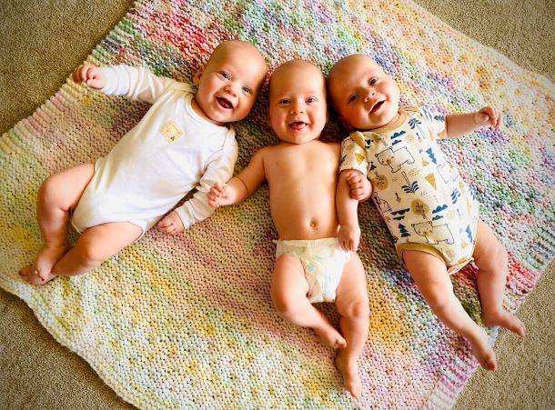 Three identical male babies smile while laying on a blanket.