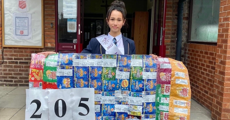 Alyssa Dean of Wales holds up blanket made from discarded chips bags as she poses outside in front of a building.