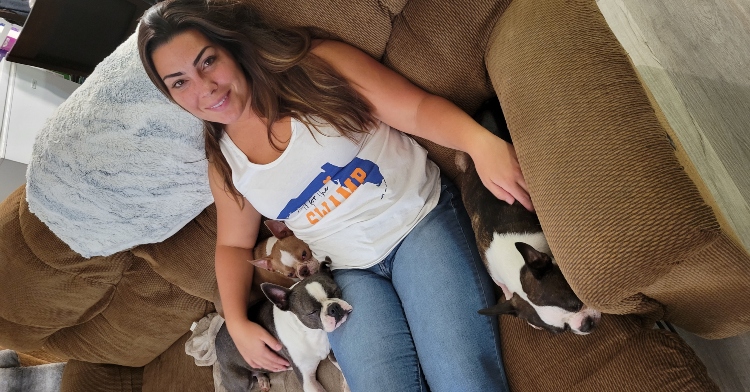 lauren sutton smiling as she sits on a couch. two of her puppies are laying next to each other, each of them resting their heads on her lap. a third puppy is laying on the other side and lauren has her hand resting on them.