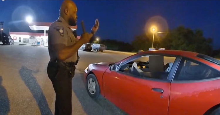 Deputy Kendrae Traylor using his hands to explain how to drive a stick shift to the young woman in the red car in front of him. The woman's face has been blurred out to protect her identity.