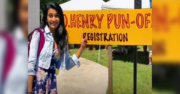 J. Maya smiling as she points to the sign behind her that reads "O. Henry Pun-Off registration."
