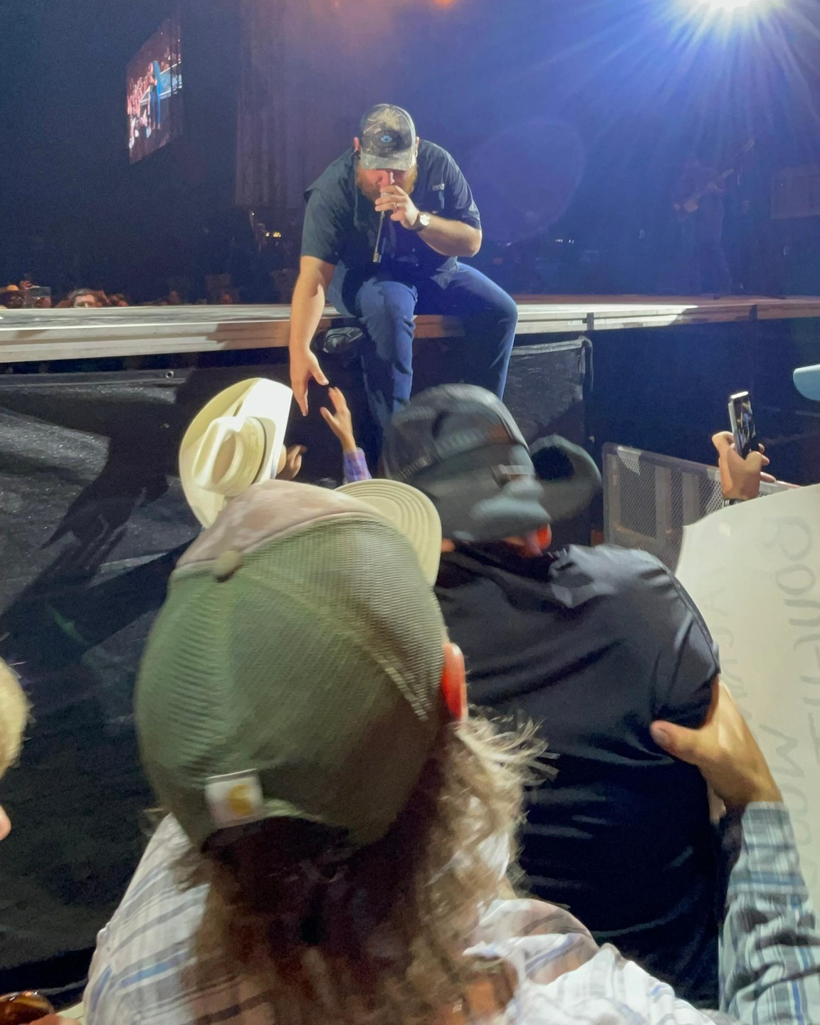 luke combs sitting on the side of the stage, legs hanging off the side. he's talking into a mic as he reaches one hand out to shake hands with tanner.
