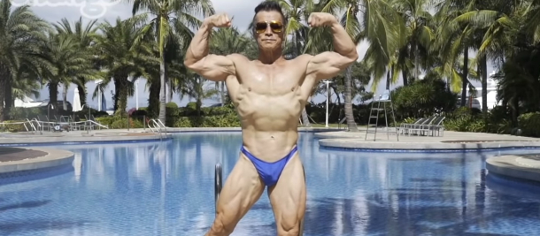 Xinmin posing near a pool. He's wearing a bathing suit and sunglasses and is showing off his impressive muscles. 