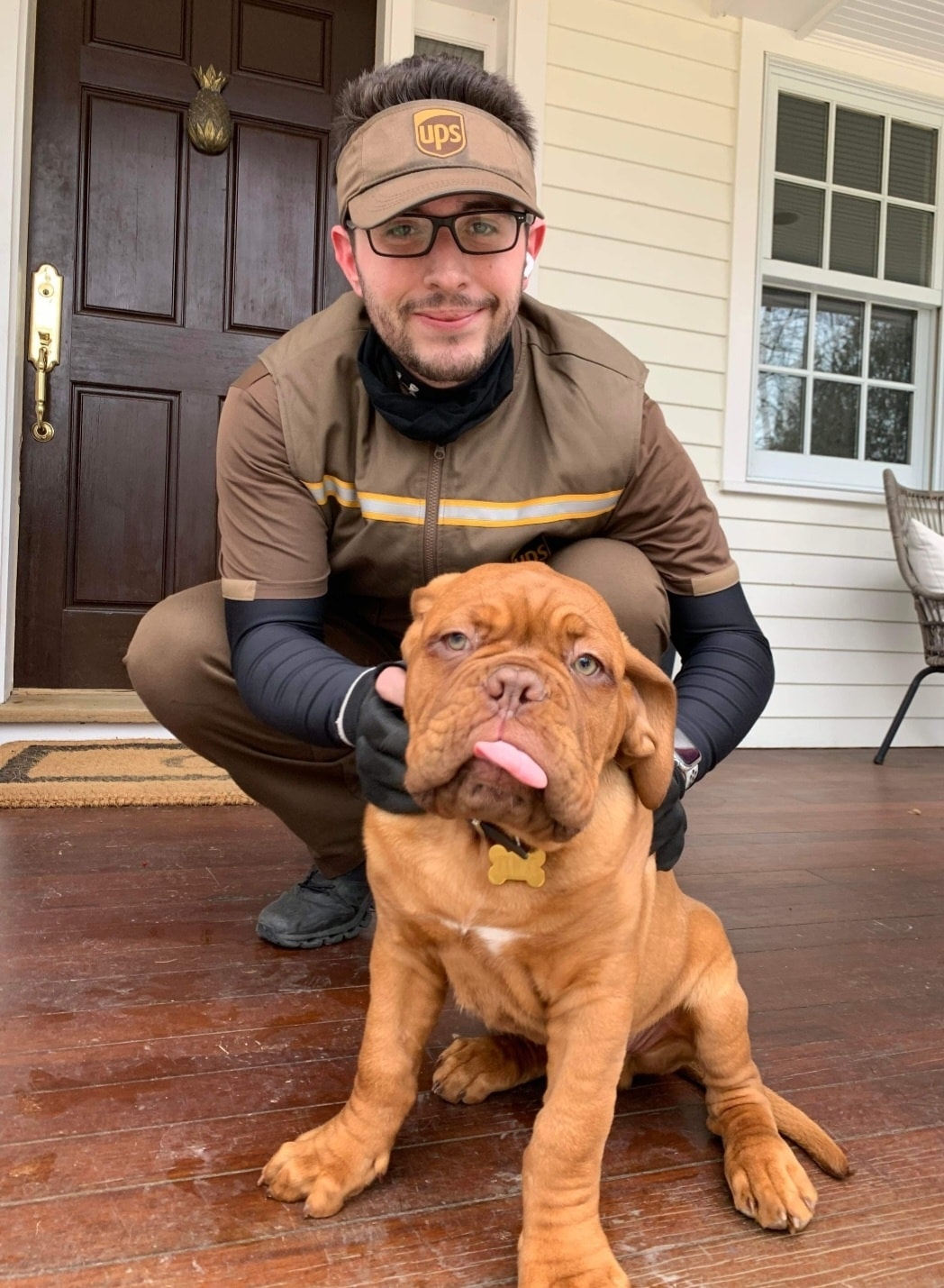 UPS driver squats on porch holding up face of puppy with his tongue out.