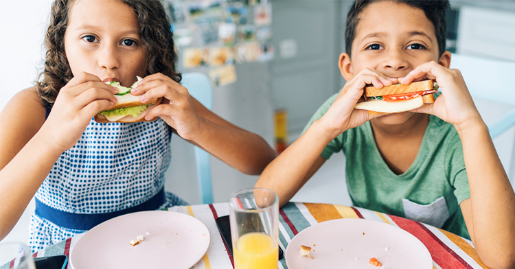 two children sitting at a table and eating sandwiches