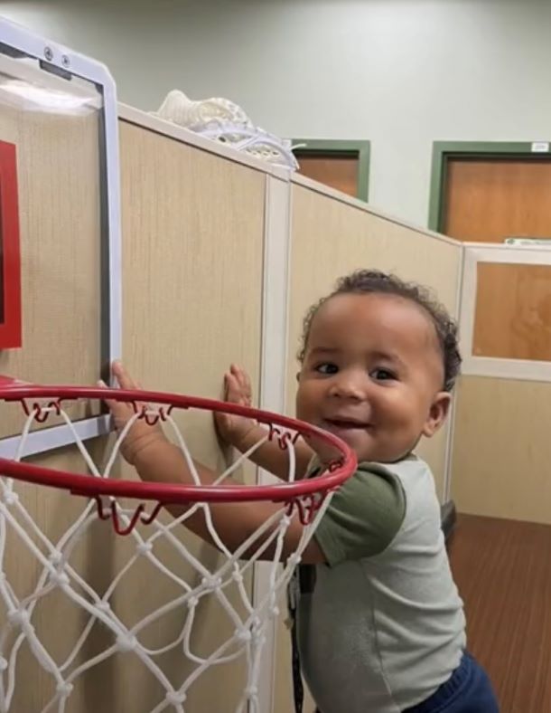 A baby boy smiling near a basketball hoop.
