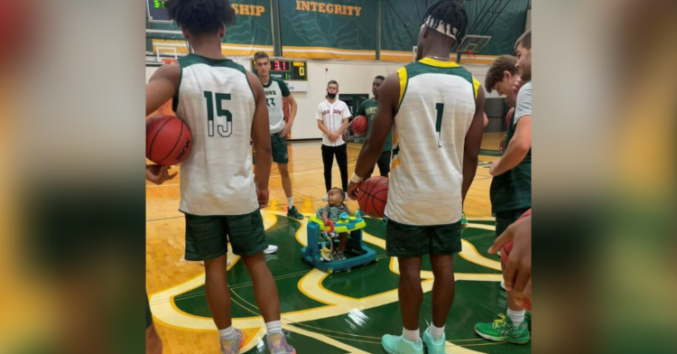 Basketball players standing around a baby sitting in a walker.