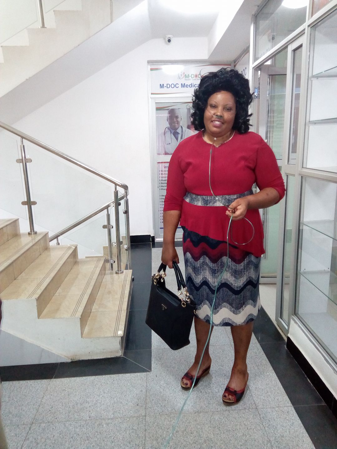 Gladys, who is dressed nicely in a top and skirt with a large purse in one hand, smiles while standing in a nice building. She is wearing an oxygen tank.