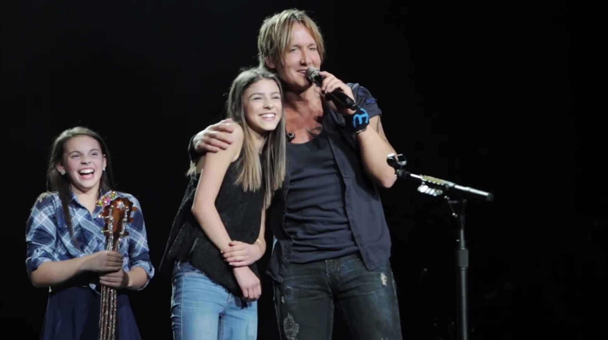 Keith Urban has his arm around 14 year old Hailey who is smiling as he talks into the mic on stage.