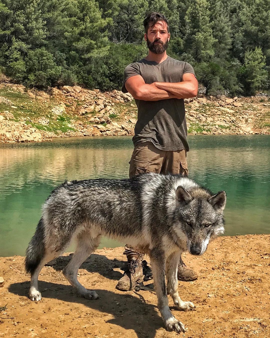 man stands with arms folded and big grey wolf hybrid at his feet