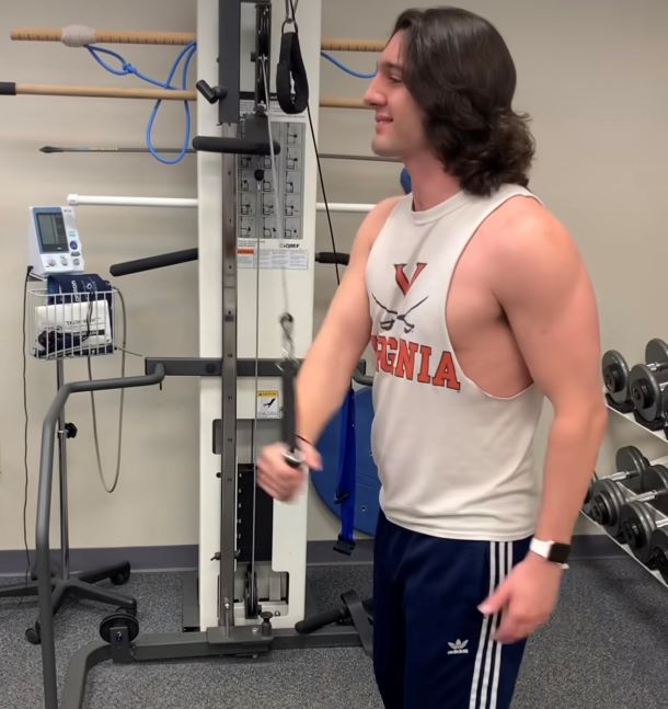 Cody smiles while exercising in the gym. He now has long hair and is much healthier looking with more prominent muscles.