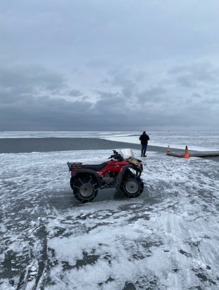 A Red Snowmobile is Parked on the Frozen Shoreline of the Lake. A Person is Standing in the Distance, Right Beside the Temporary Bridge.  