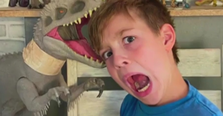 Little boy with his mouth wide open, as if screaming, being silly as his toy dinosaur "bites" his head.