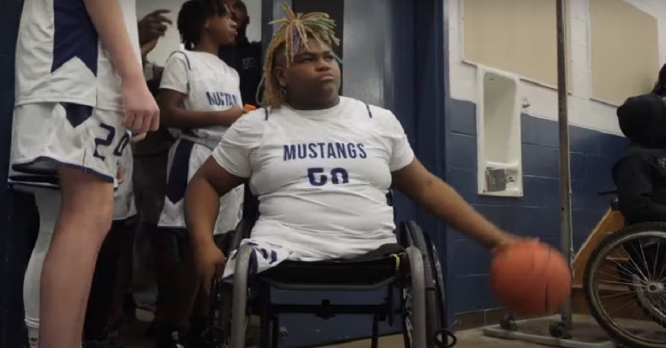 Teen boy with no legs sitting in a wheelchair, dribbling a basketball while wearing a basketball uniform