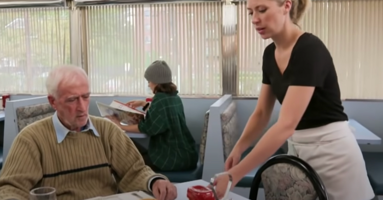 an old man sitting at a restaurant table by himself while a waitress is standing and taking his order.