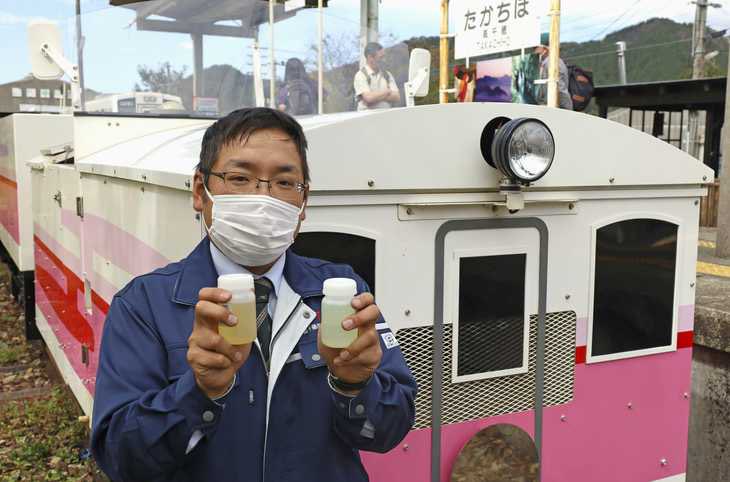 Man wearing a face mask holds up two vials of biodiesel used to power the Amaterasu Railway. The pink and white train is parked just behind him.