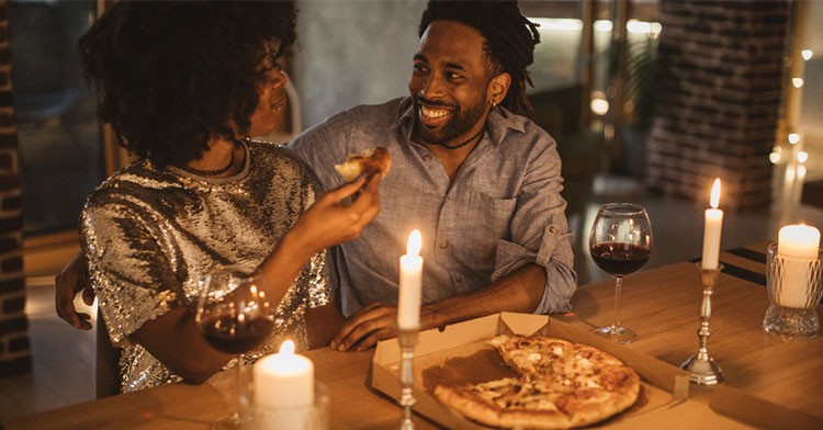 man and woman smiling at eat other and eating a candlelit pizza dinner