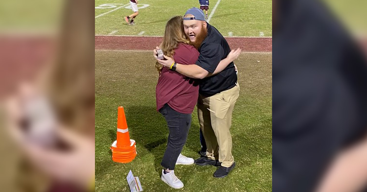 Man showing off an engagement ring to a crowd as he hugs his girlfriend who has no idea he has a ring.