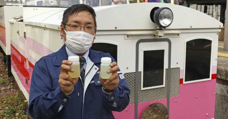 Man wearing a face mask holds up two vials of biodiesel used to power the Amaterasu Railway. The pink and white train is parked just behind him.