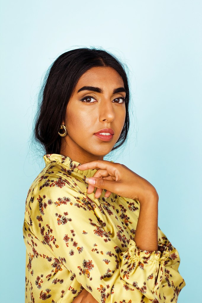 Poet Rupi Kaur posing in a yellow silk shirt covered in flowers standing in front of a blue background 