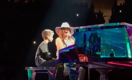 Lady Gaga playing the piano and singing to Owen, who sits beside her on the piano bench.