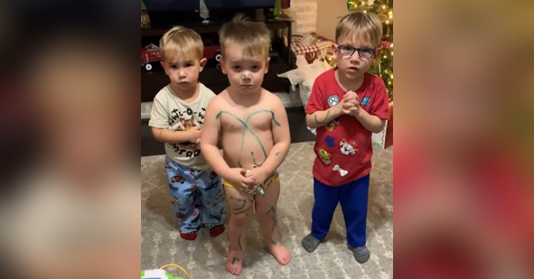 Three toddlers stand in the middle of a room and look guilty, one of them is just wearing his underwear and has marker all over him.
