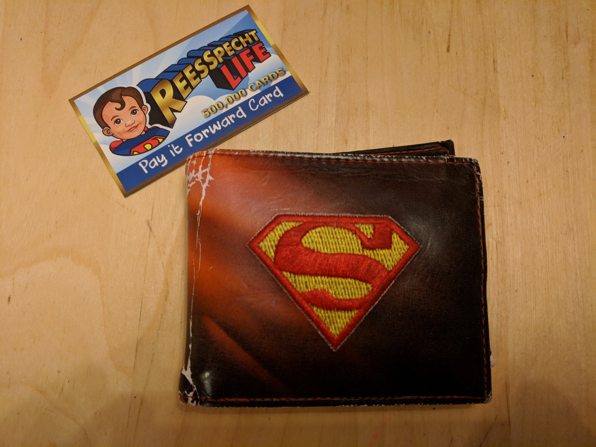 a car that reads "ReeSpecht Life 500,000 cards, pay it forward card," with a cartoon image of Rees on it, sitting next to a beat-up Superman wallet.