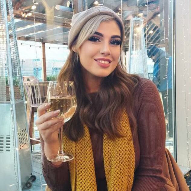 Daisy wearing a long yellow scarf and holding a glass of white wine.