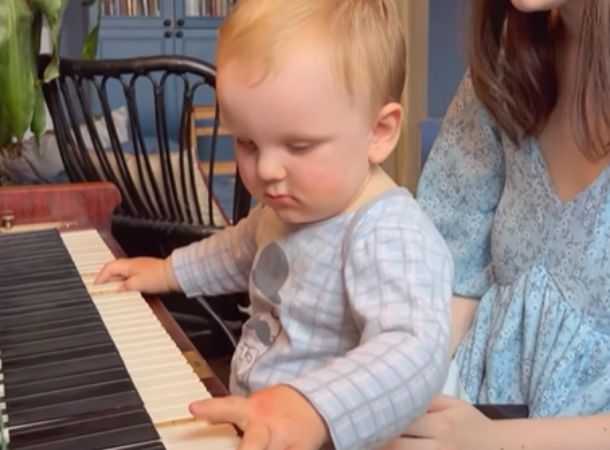 An adorable one-year-old plays the piano on his sibling's lap.