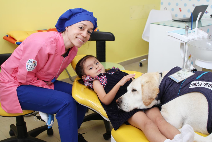 A dental worker at Parque Dental smiles next to a little girl who is laying in a dental chair. Aldo the dog lays next to the girl, resting his head on her lap. His eyes are closed and he looks calm.