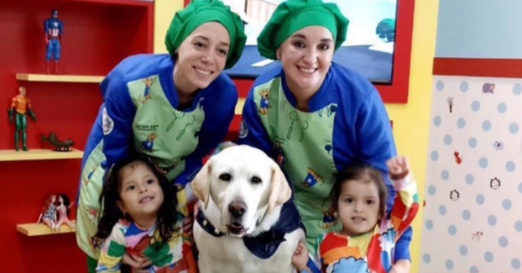 Two dental workers at Parque Dental smile and pose with two young patients and Aldo the dog. The room they are in is colorful and has bean bag chairs and a shelf with action figures and dolls.