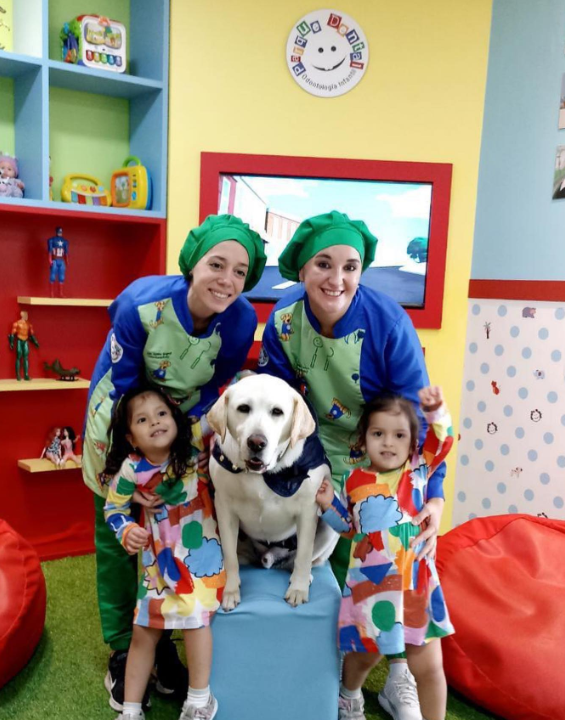 Two dental workers at Parque Dental smile and pose with two young patients and Aldo the dog. The room they are in is colorful and has bean bag chairs and a shelf with action figures and dolls.
